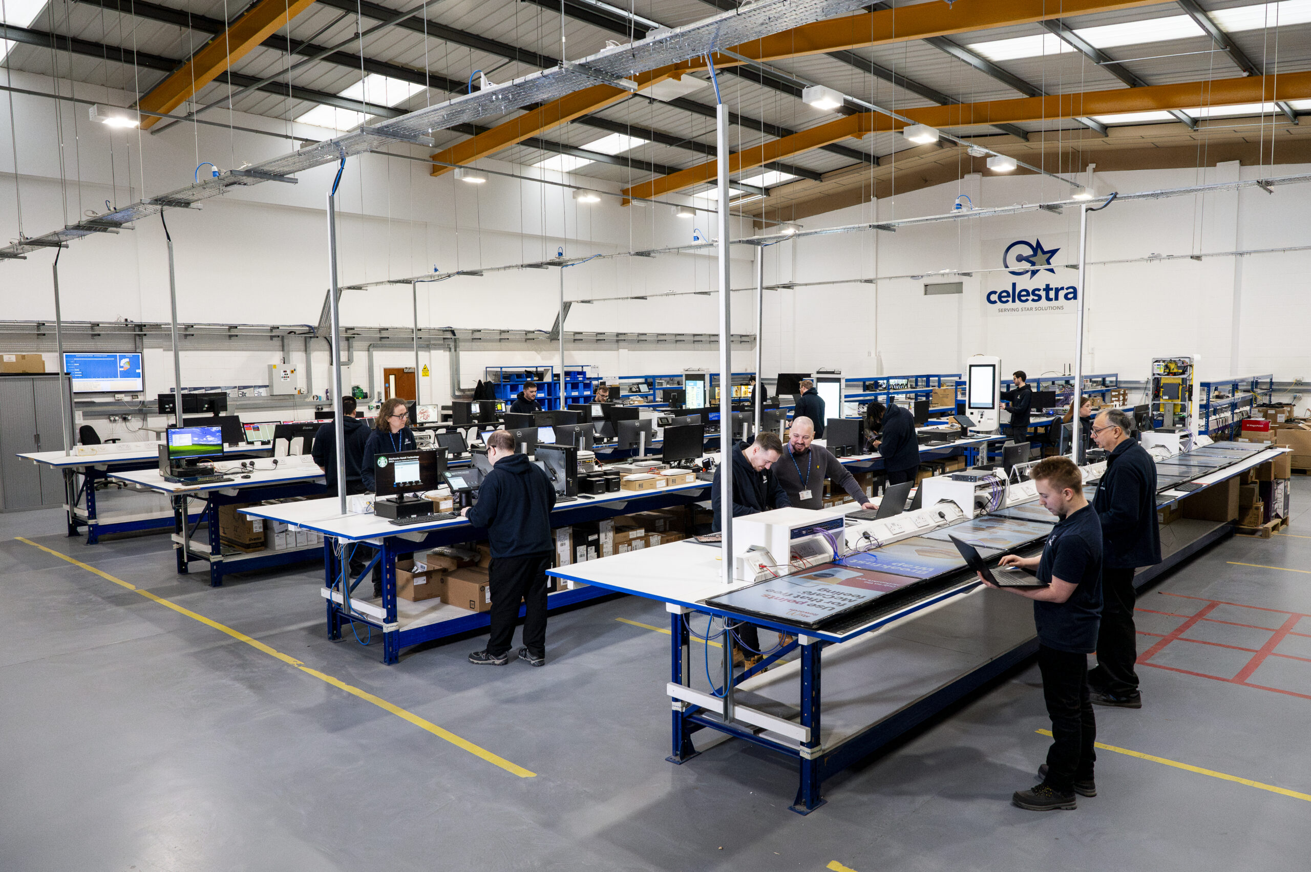 A configuration centre. There are multiple people working at waist high level work benches. They are configuring various Starbucks Tills, EPoS technology, McDoanlds Kiosks, and Digital Media Screens ready for installation onto site. the Celestra logo is centre placed in the image showing the C-Star brand.
