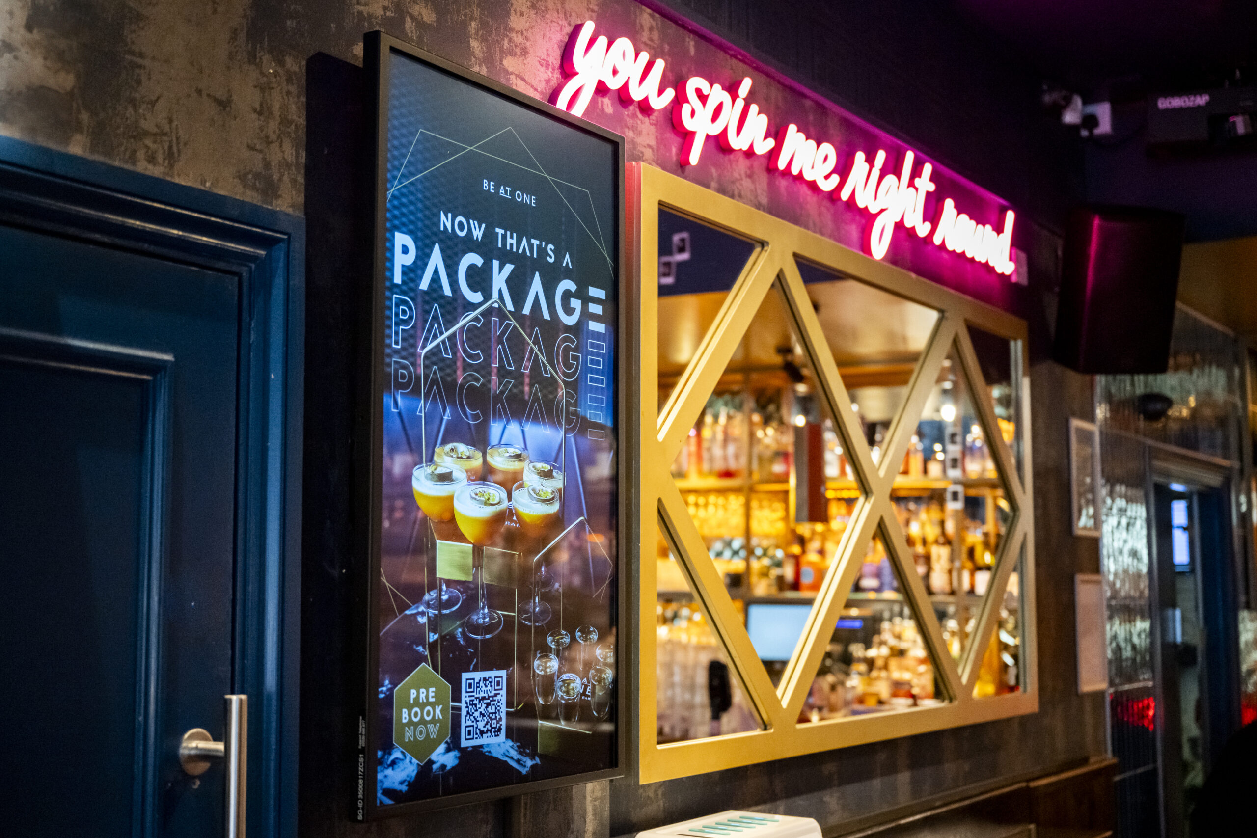 The image is of a wall in a Stongate Be At One venue. On the wall there is a fully fucntioning Digital Media Screen displaying and advert for booking a package deal using the venues app. The rest of the image is made up of a mirror with gold lattice work across the front and a bright neon pink sign across the top which reads 'you spin me right round'.