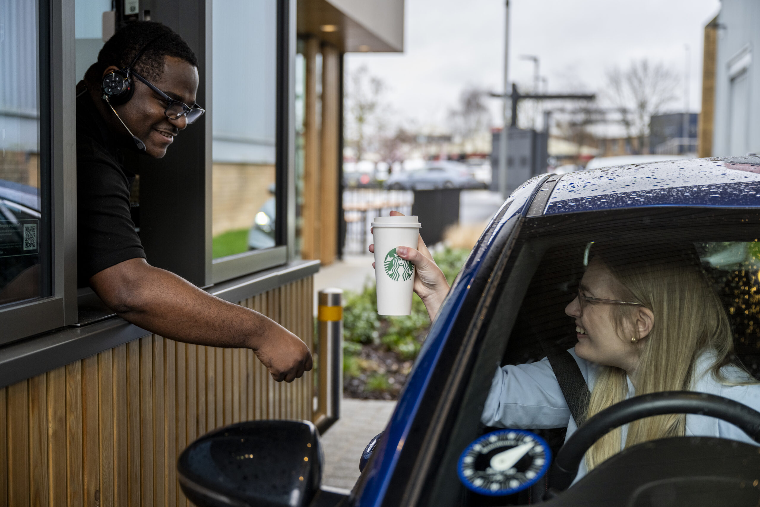 A man is handing a Starbucks Coffee out the kisok window to a lady as she sits in the Drive Thru lane in a blue car.