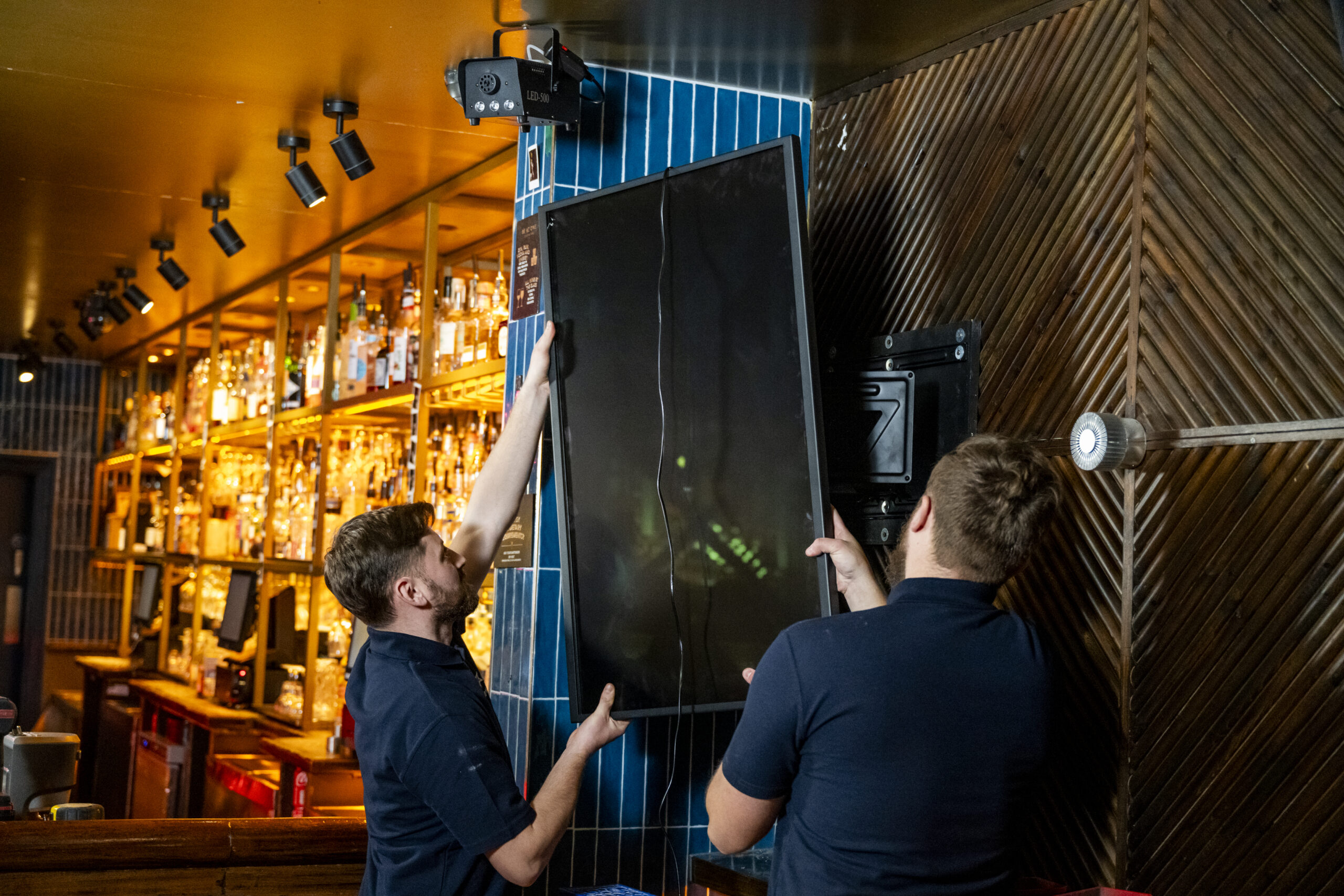 Two installation engineers lifting a digital screen up on to a wall mounted bracket in a Stonegate Be At One venue.